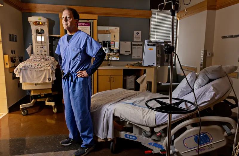 A white man in scrubs stands in a hospital room.