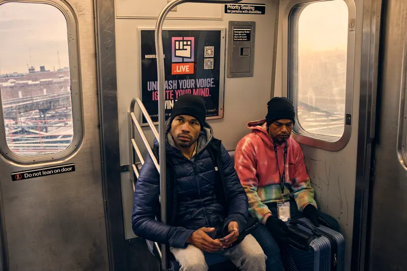 Two Hispanic men sit together on a subway car. Larez holds a suitcase between his knees.