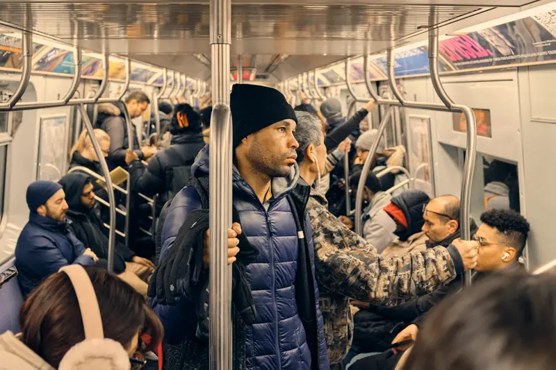 A Hispanic man in a blue coat stands in a crowded subway car holding a pole.