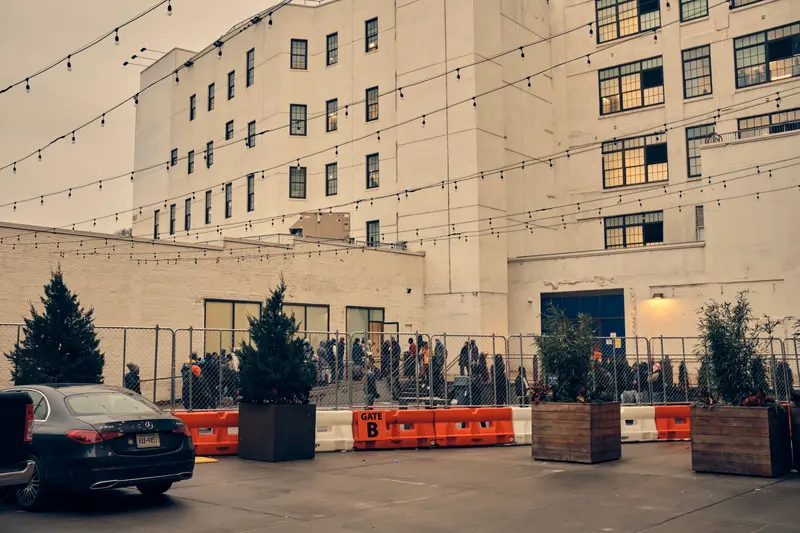 A long line of people in winter clothes wait in a fenced-off area of a parking lot.