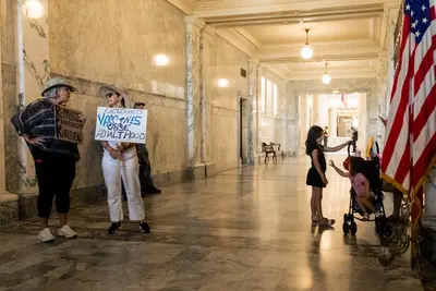 Inside a long marble hallway, two little girls play near an American flag. Across the hallway, two female protesters look at one another. One holds a sign that reads: “Reprehensible, Fraudulent Knucklehead.” The other woman holds a sign reading “Childhood vaccines cause adulthood.”