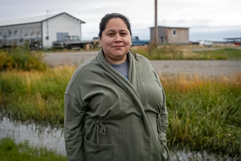 A woman in a green jacket stands amid reeds in front of a low white building.