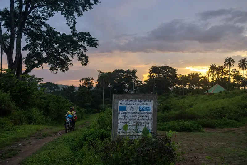 Two people on a motorbike ride by a worn down sign as the sun sets over the nearby forest.