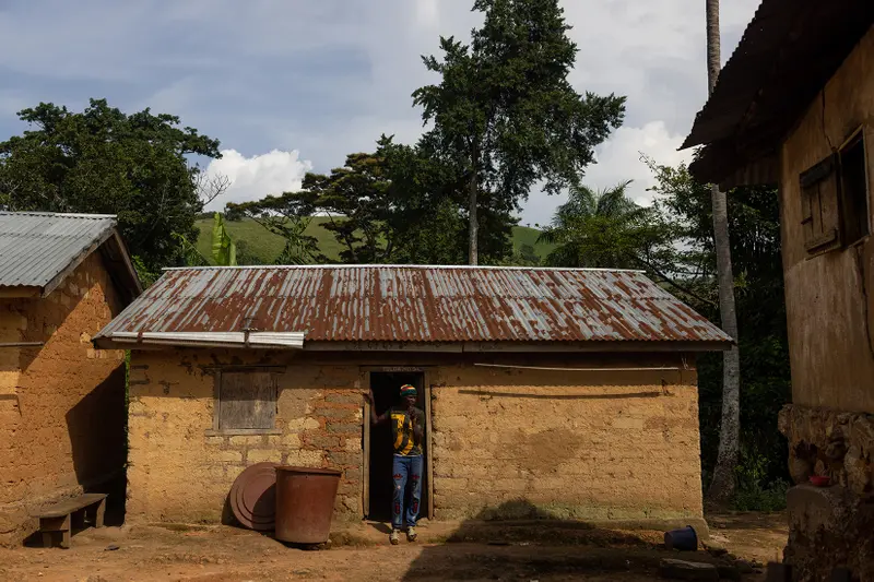 A man stands in the doorway of a yellow brick house with a rusted tin roof. Trees tower over the building behind him.