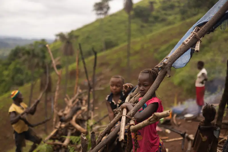 A girl in a pink shirt stands next to a wooden structure, staring out at the landscape. She is surrounded by other children and her father in the background, who is working.