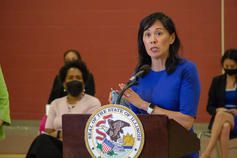 An Asian woman in a blue dress stands behind a lectern bearing the seal of the state of Illinois