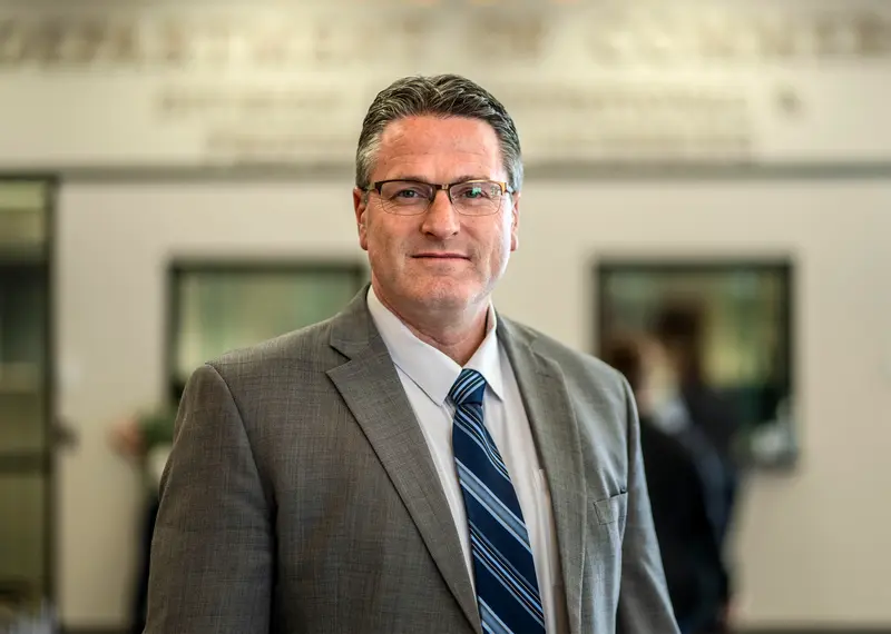A man with salt-and-pepper hair wearing a grey suit and blue striped tie.
