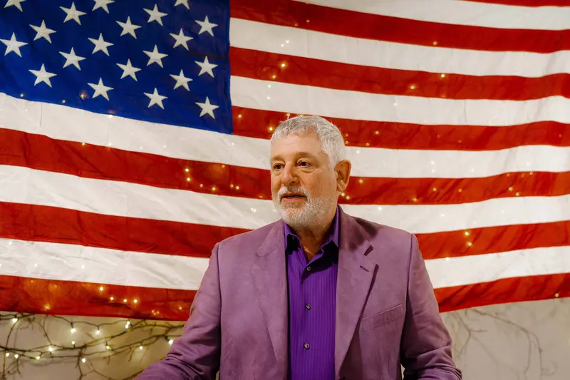 A head-and-shoulders shot of a white man with silver hair and a beard stands in front of a large American flag. He's wearing a purple shirt and blazer.