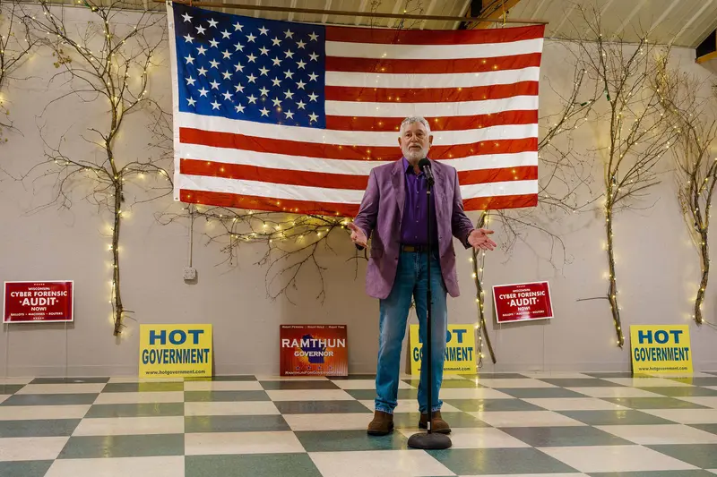 A full-length shot of Jay Stone in front of a microphone, with yard signs for H.O.T. Government, a "cyber forensic audit" and Ramthun for governor in the background.
