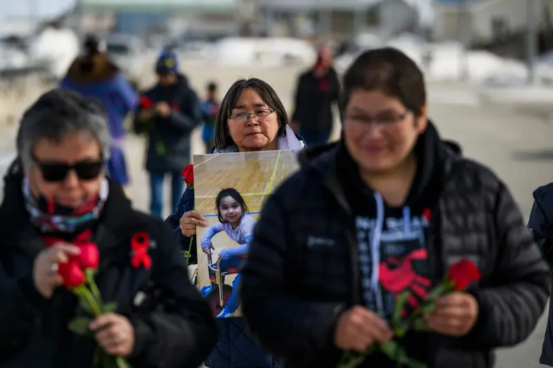 A group of people wearing coats and jackets hold red roses. A woman with chin length hair and bangs looks into the camera and holds a blown-up photo of a little girl on a scooter.