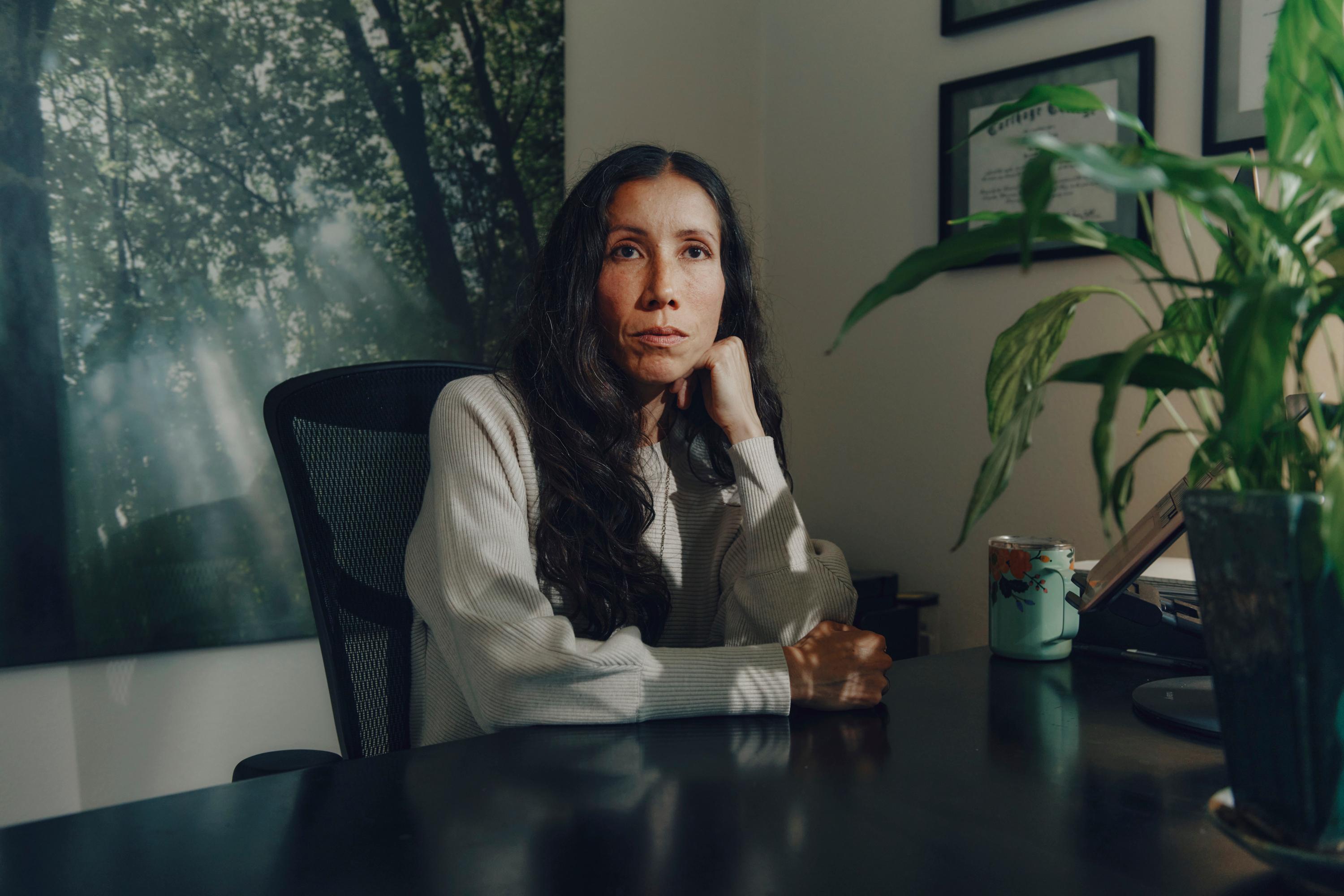 A woman sits in her office leaning on her hand while she looks above the camera. There is artwork of a forest behind her.