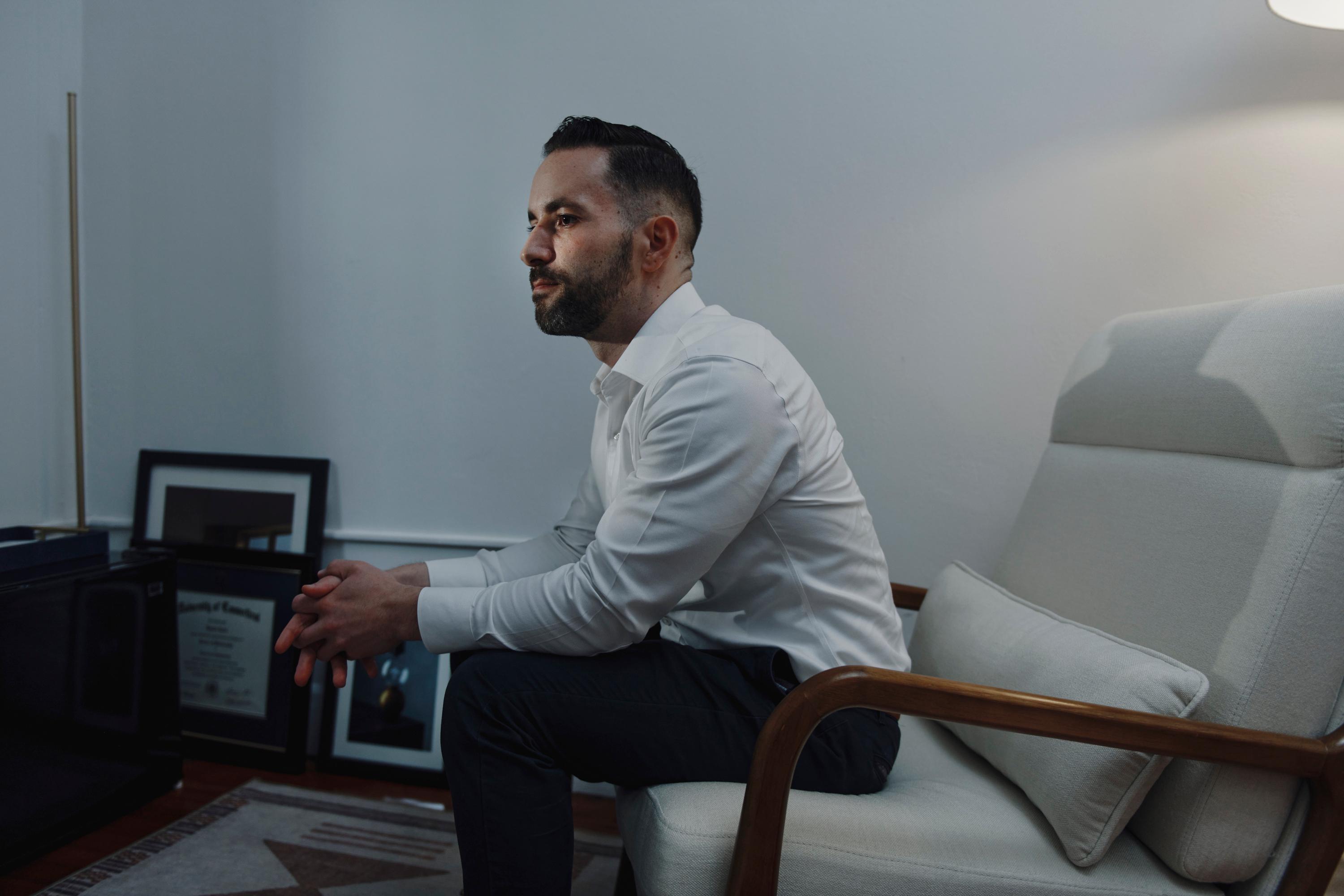 A man sits in a white chair in his office. His hands are clasped together and he’s looking off into the distance. 