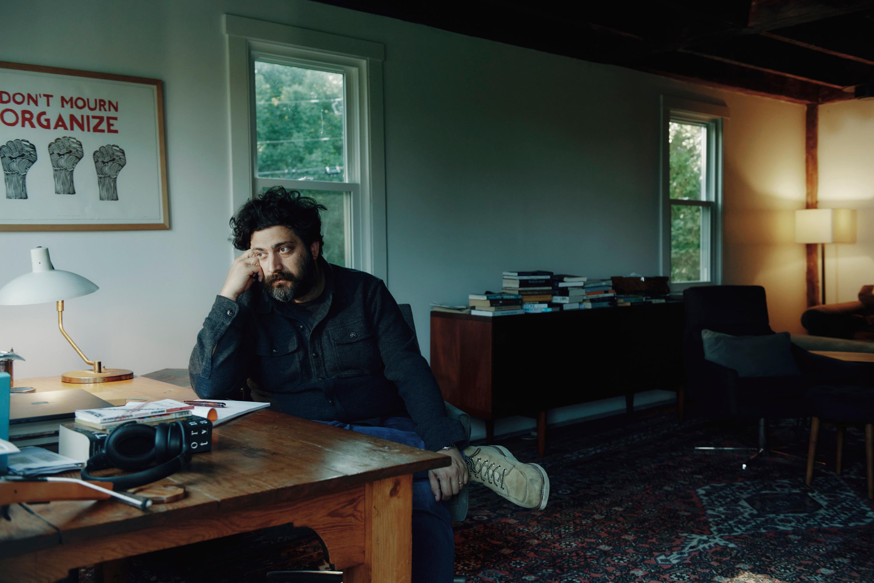 A man sits at a desk in his office while he looks out the window. A lamp shines a warm light in the background.