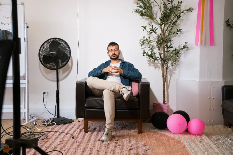 A man wearing khakis, a blue jacket and white sneakers sits in a leather armchair in an office in a renovated loft, decorated with deflated pink balloons, some paper streamers and a potted artificial tree.