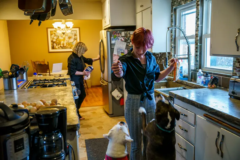 Two dogs jump toward Blake in a kitchen with concrete countertops while in the background Jennifer reaches for the fridge.